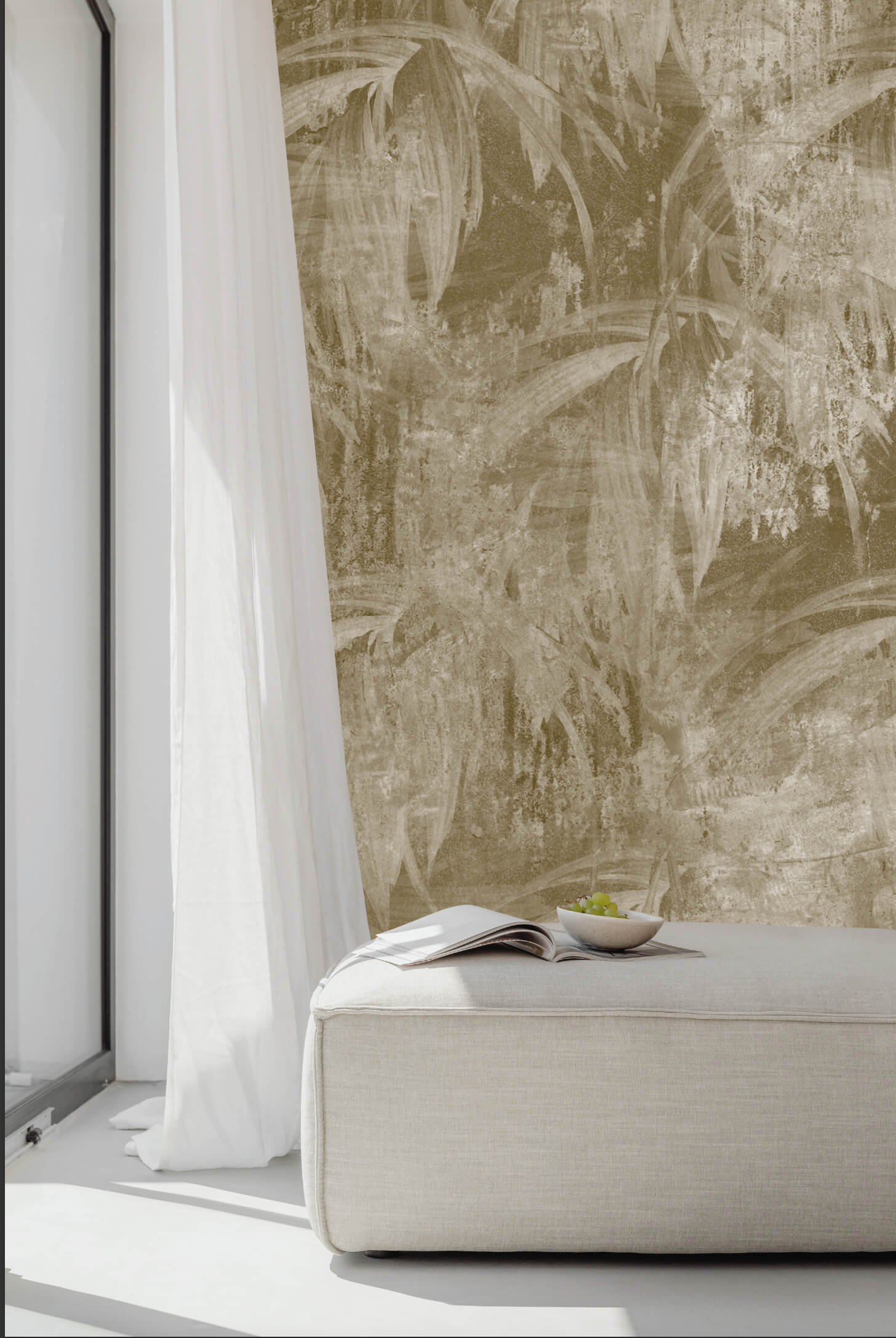 Dettaglio di un interno luminoso con panca imbottita in tessuto chiaro e una tenda bianca accanto a una finestra. La parete è rivestita con un murale astratto a tema foglie di palma in tonalità sabbia.